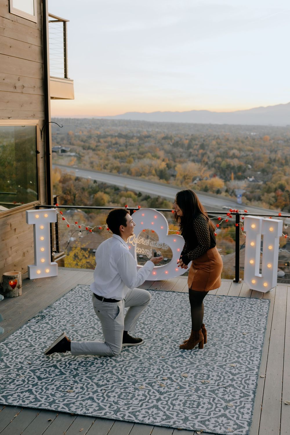 Two people enjoying a scenic mountain view from a modern balcony with string lights at sunset.