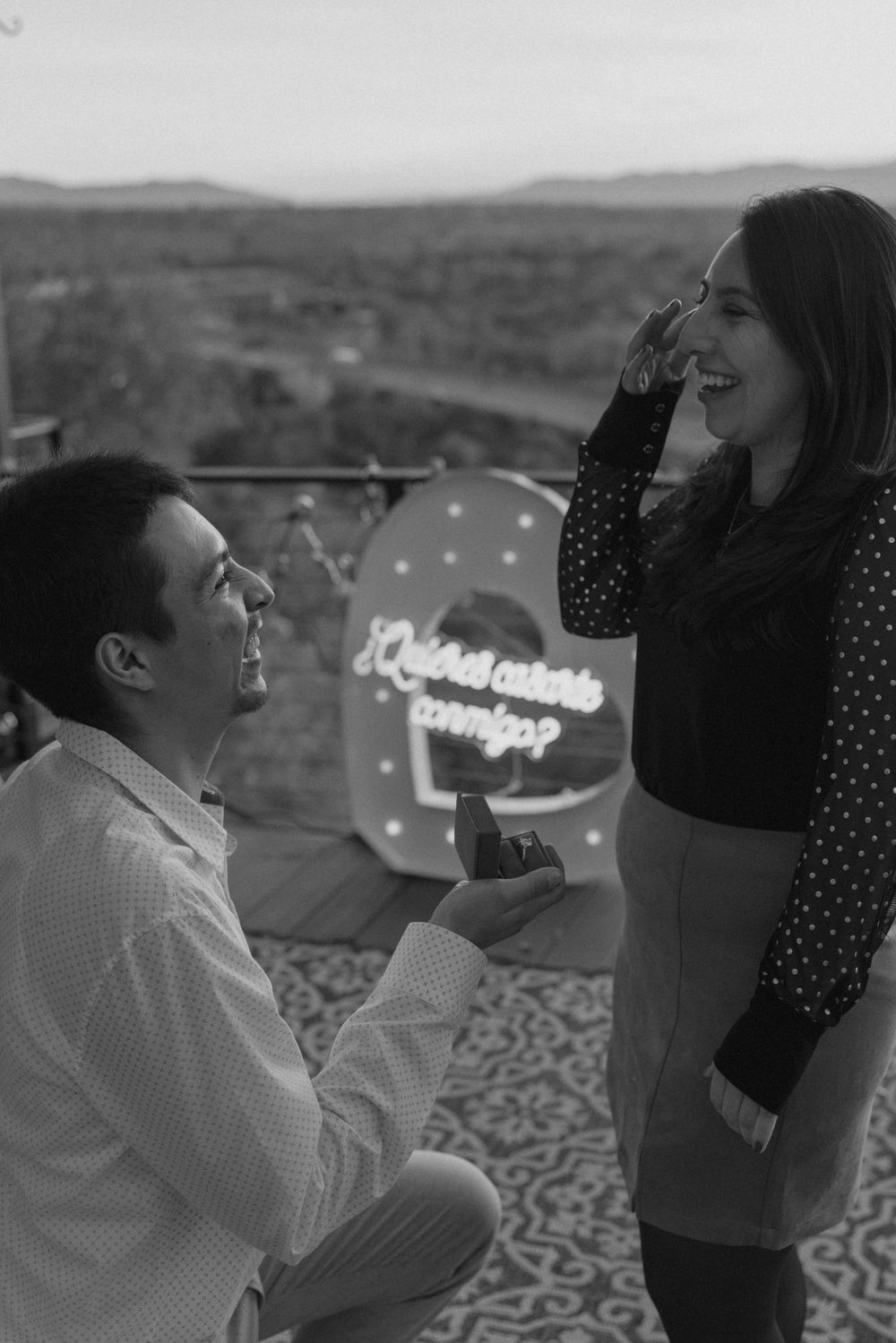 A romantic proposal scene unfolds on a balcony with city lights and mountains in the background.