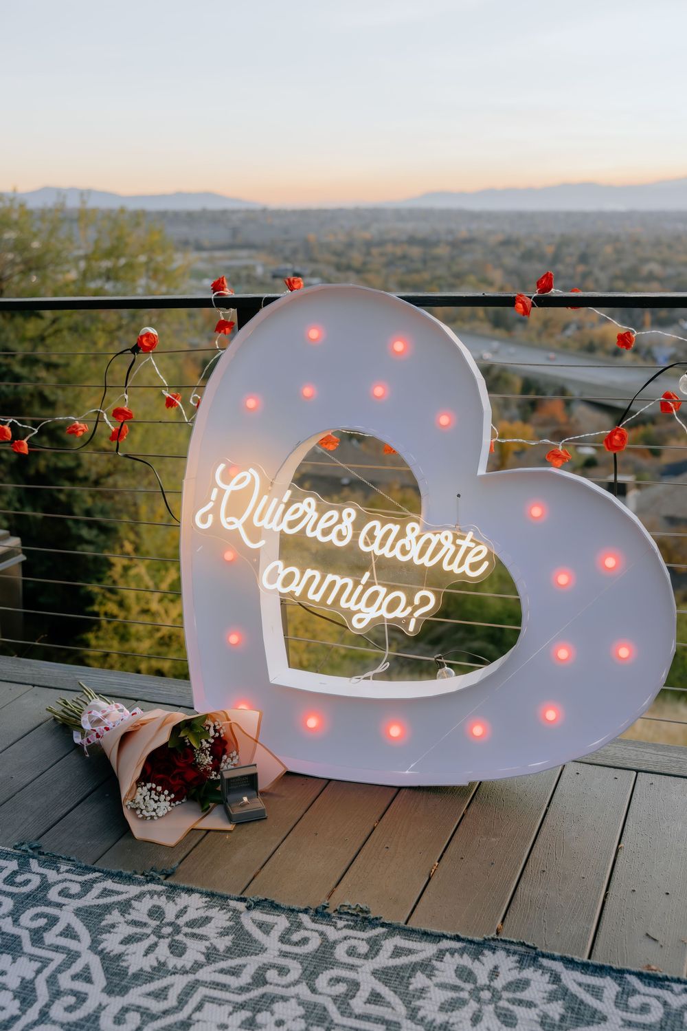 Large illuminated letters spelling out a proposal message on a wooden deck at sunset with mountain views in background.