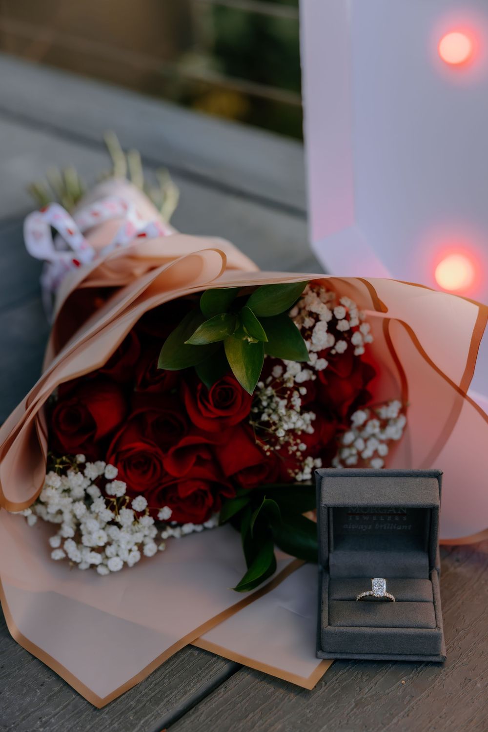 A romantic proposal setup with red roses and baby's breath flowers next to an open ring box with soft pink lighting.