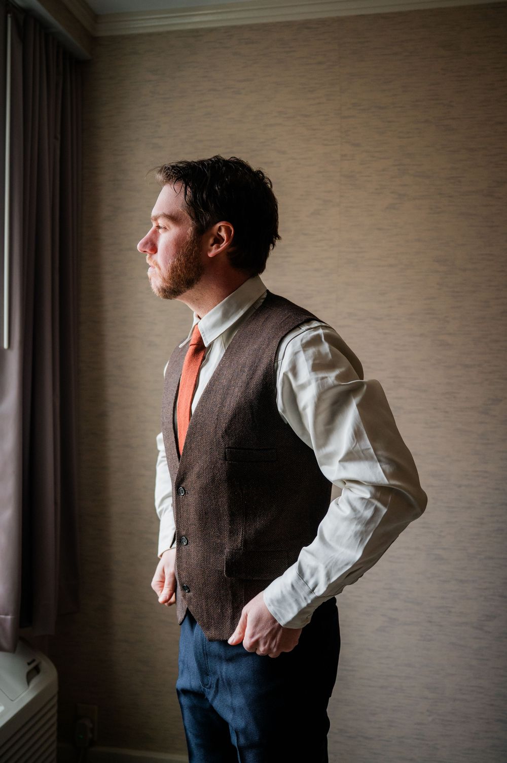 A person in business attire gazes thoughtfully out a window while wearing a brown vest and orange tie.