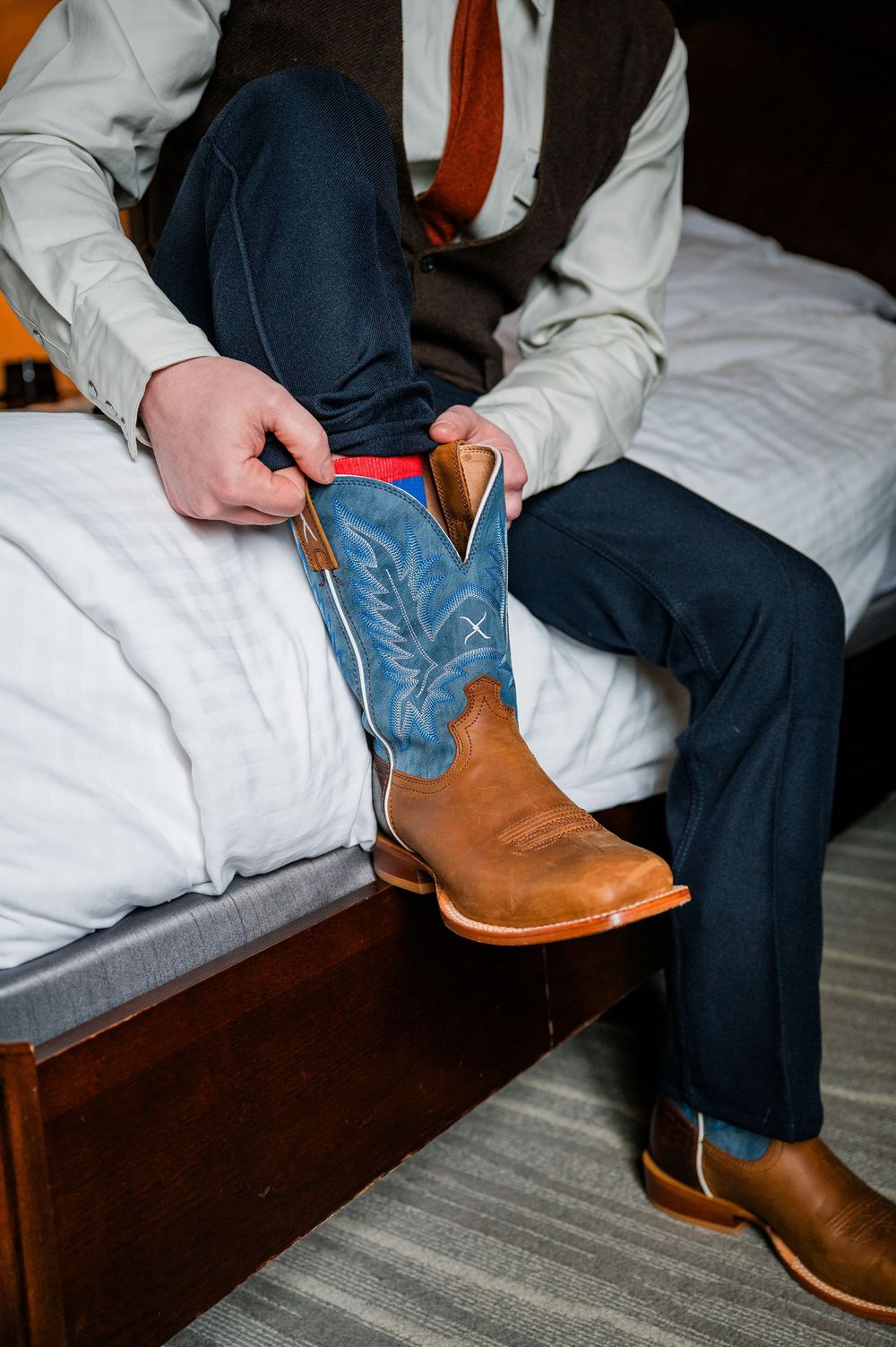 Detail shots of blue and brown cowboy boots being put on while sitting on bed edge.