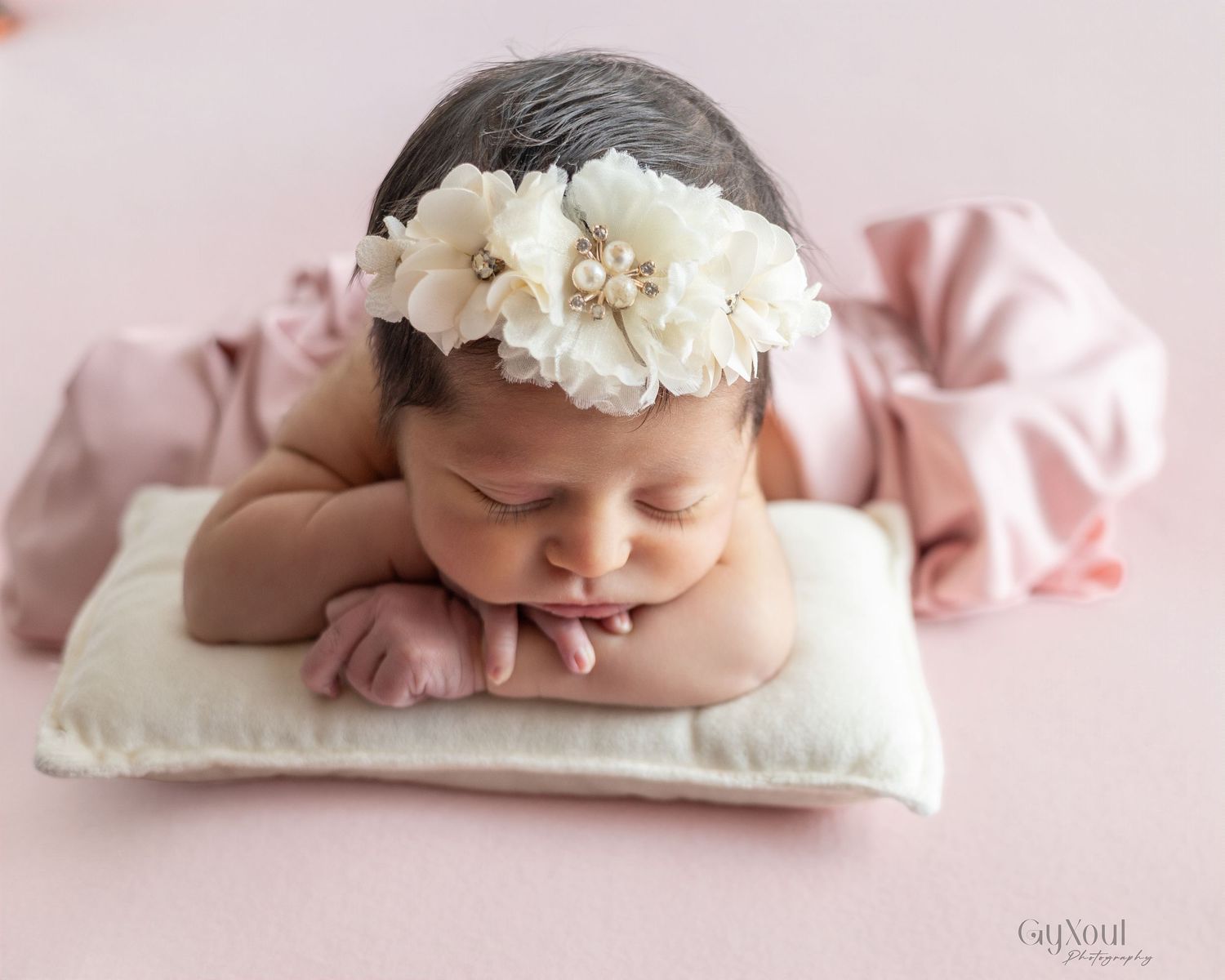 A sleeping newborn baby wearing a white floral headband lies peacefully on a pink blanket in a studio setting.