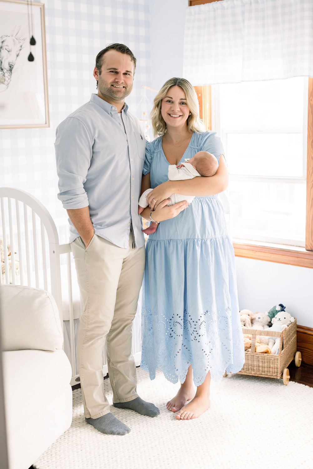 A couple in light blue and beige outfits poses together with a newborn baby in a bright sunlit room.