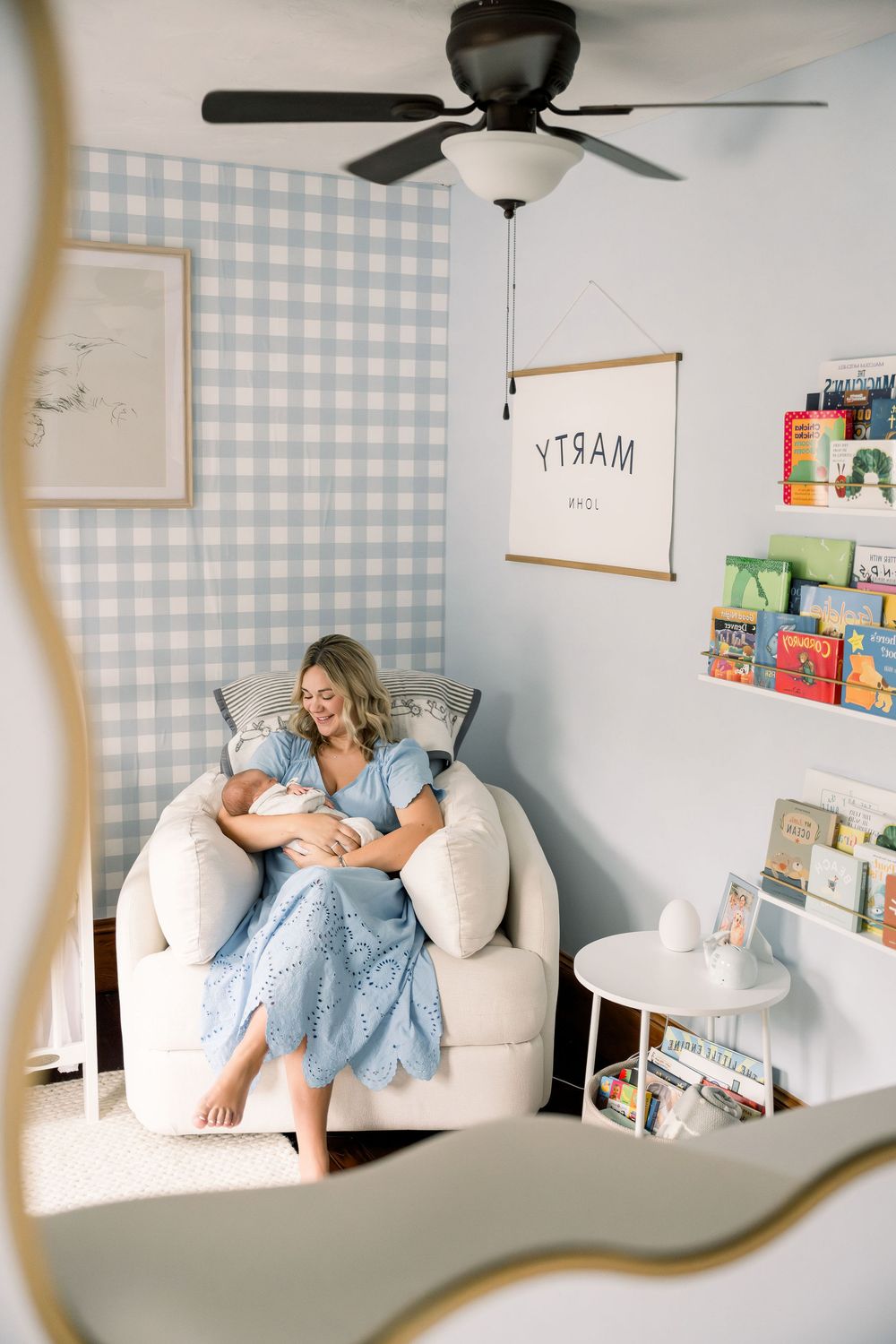 Cozy nursery corner with blue gingham wallpaper, white armchair, and floating bookshelves displaying children's books.