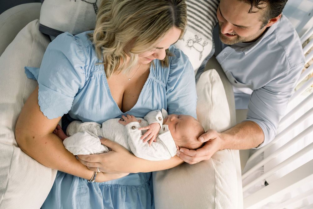 Parents in light blue and white clothing tenderly hold their newborn baby while resting on white bedding.