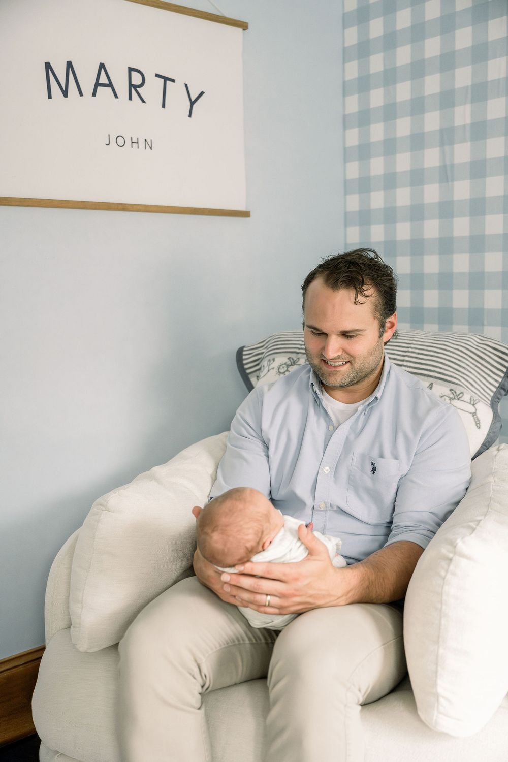 A person in a light blue shirt sits on a cream-colored couch holding a baby against a blue and white checkered wall.