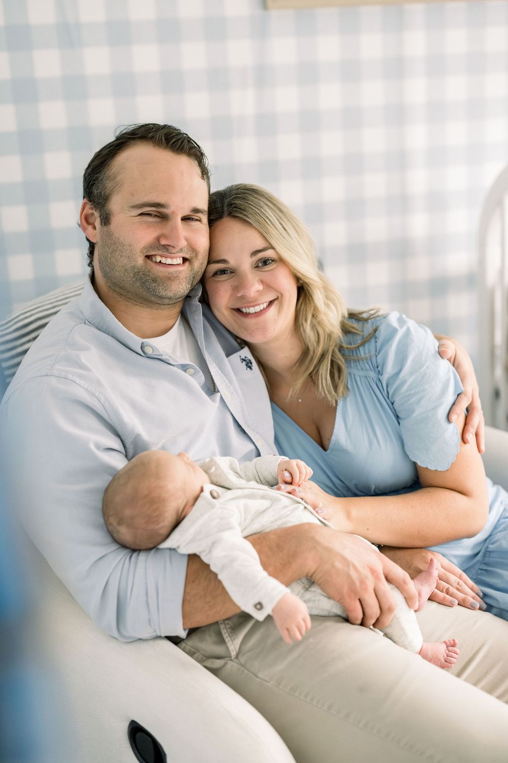 A couple sits together on a couch holding their sleeping newborn baby in a bright, airy room with checkered backdrop.