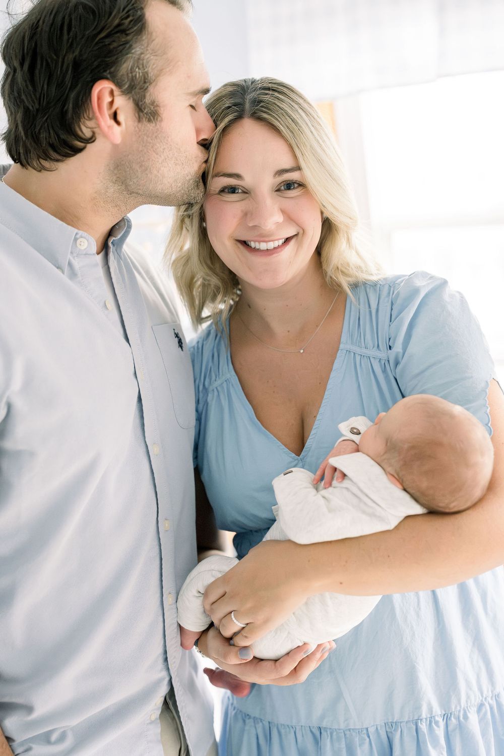 A couple shares an intimate moment while holding their newborn baby, with the mother wearing a light blue dress.