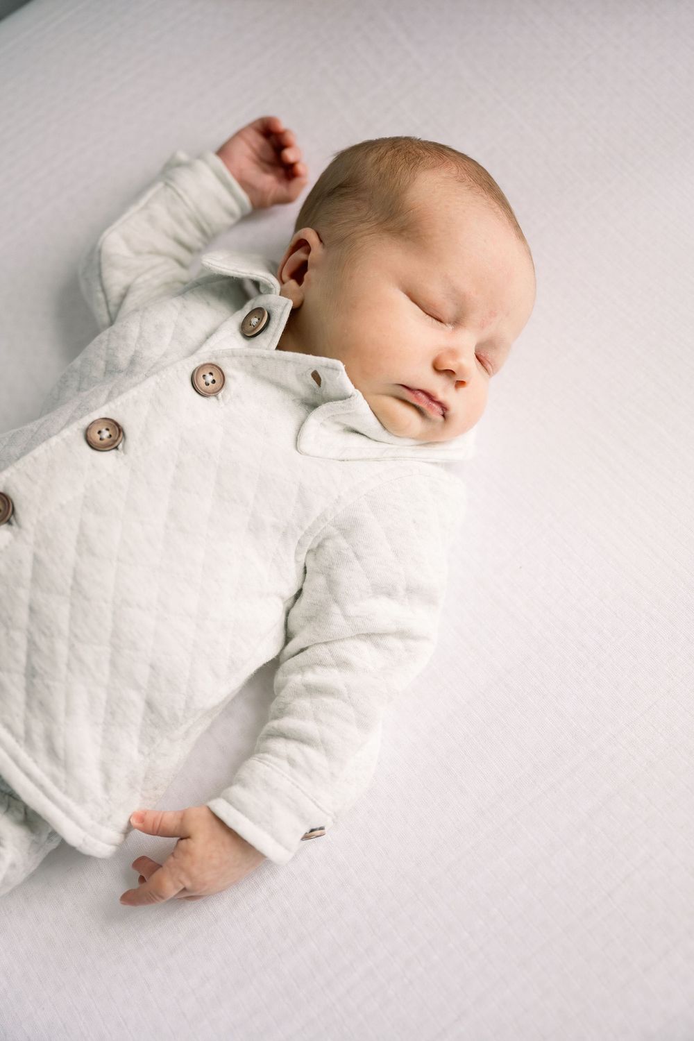 A sleeping baby in white quilted clothing lies peacefully on a light background.