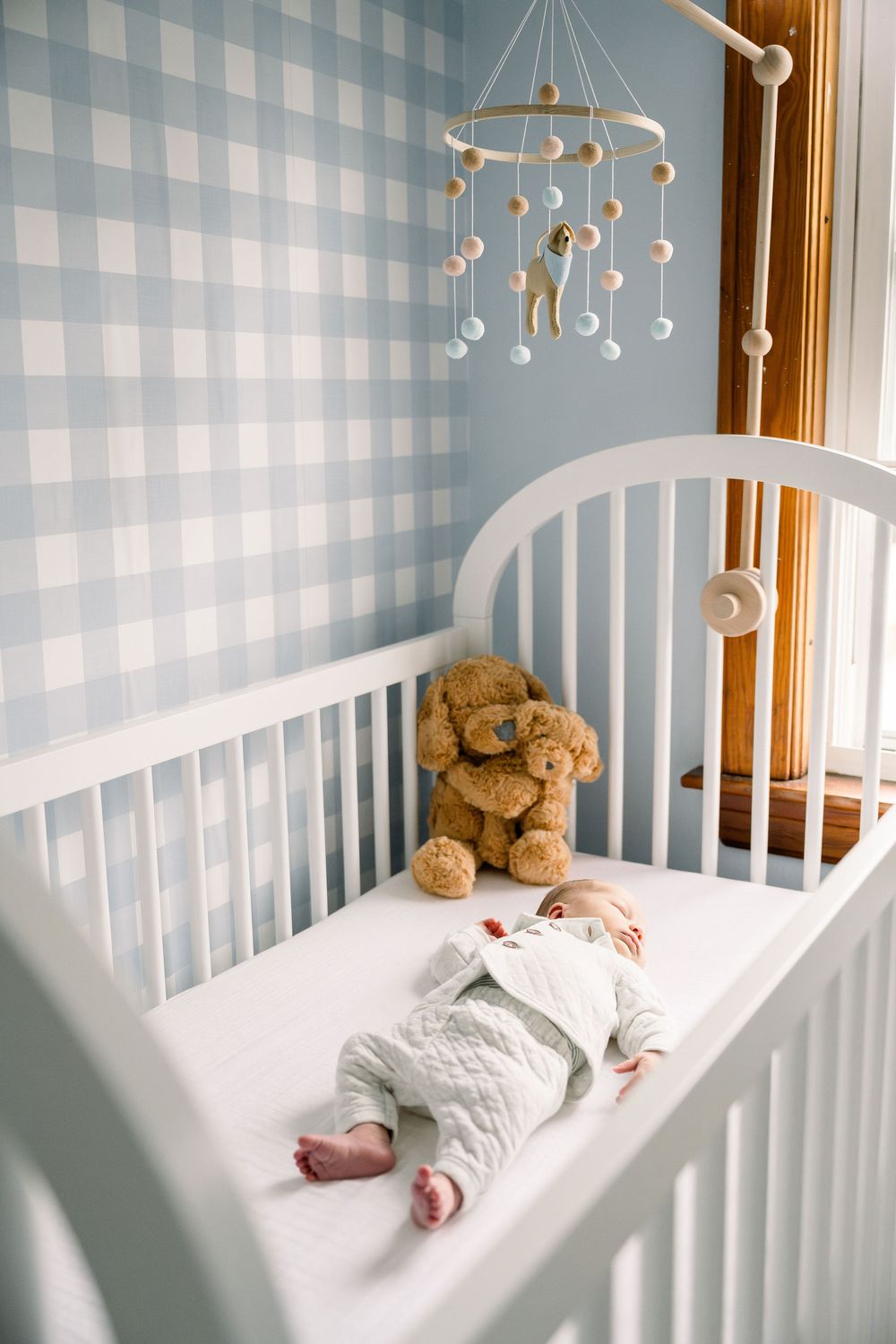 A white crib with a teddy bear and mobile featuring a blue gingham wallpaper background in a nursery setting.