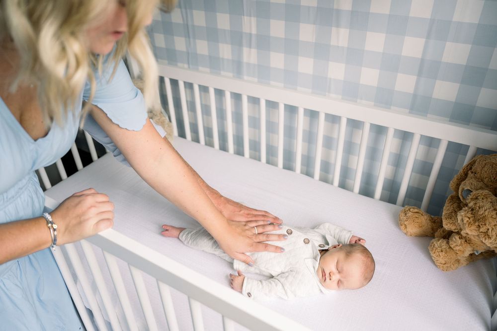Parent gently placing newborn baby into white crib with soft bedding and plush teddy bear nearby.