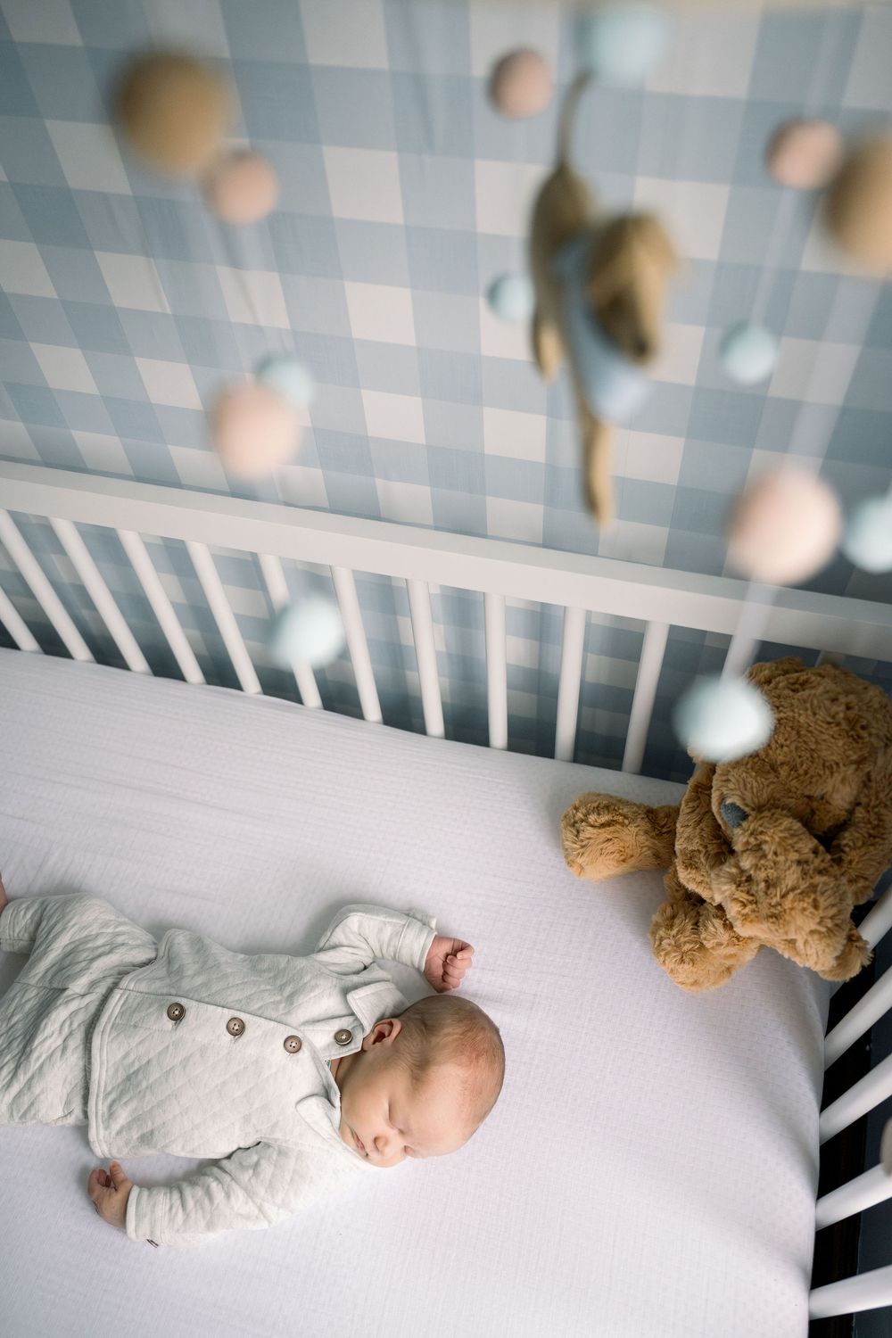 A sleeping baby in gray clothing rests in a white crib with a teddy bear.