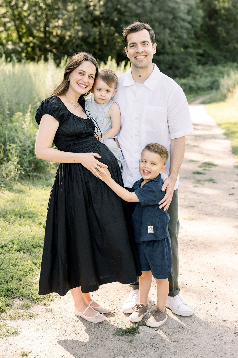 Family in coordinating black and white outfits poses together on a sunlit dirt path in a natural outdoor setting.