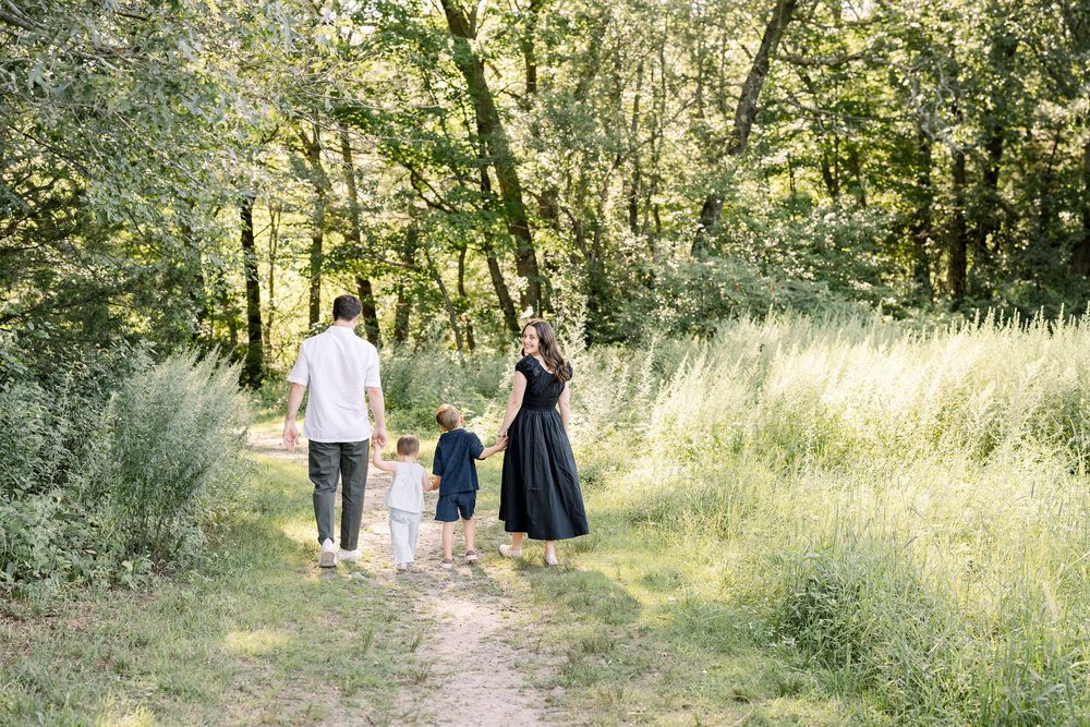 People walking along a scenic woodland path surrounded by tall grass and trees in dappled sunlight.