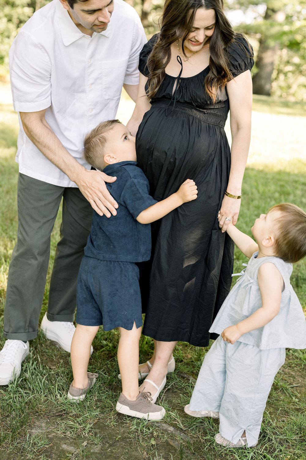 A family stands together outdoors, with a pregnant woman in a black dress and two young children hugging her.