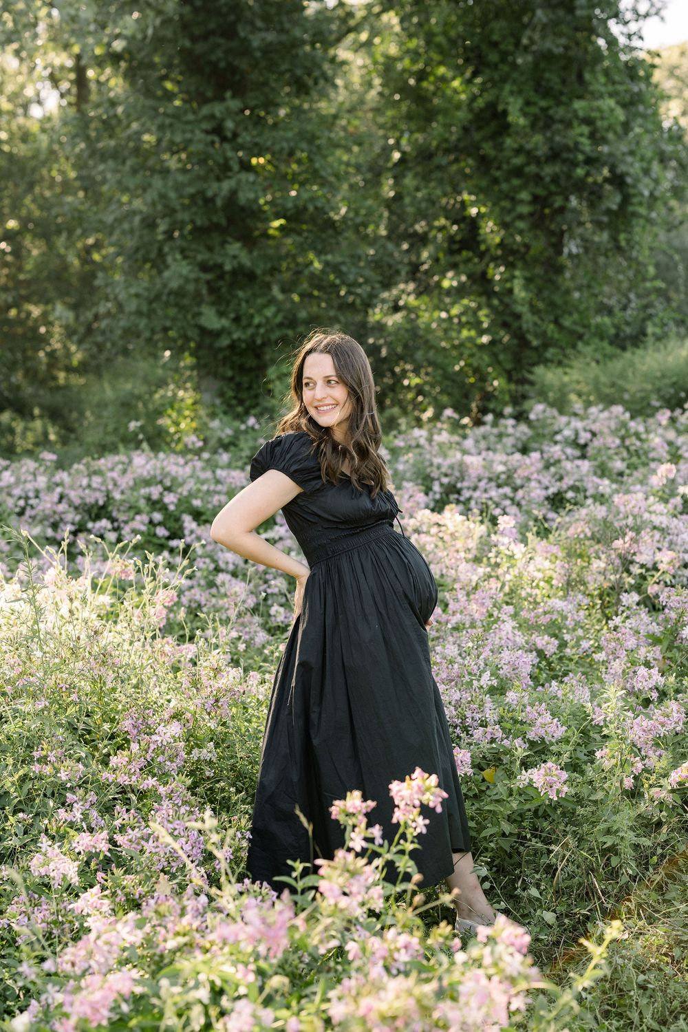 Woman in black dress twirls through a field of pink wildflowers during a sunny outdoor photoshoot.