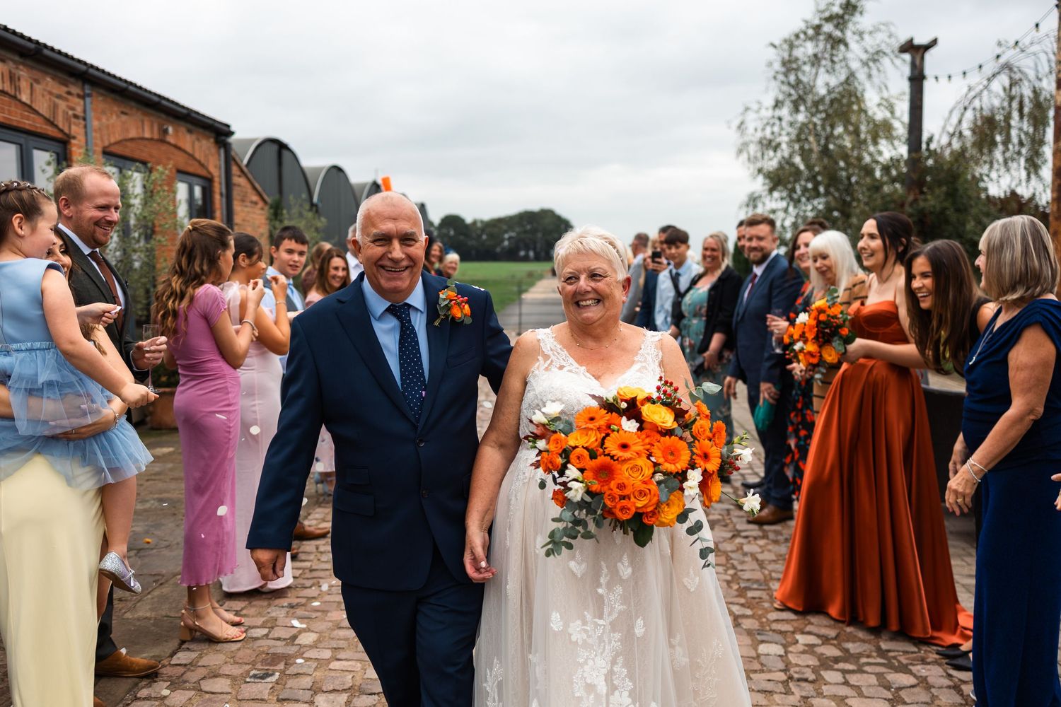 Wedding party walks down brick pathway with orange and rust colored autumn wedding decor and attire.