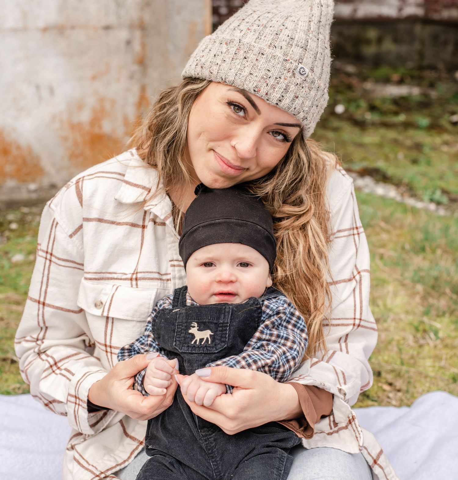 A parent and baby sit together on a blanket outdoors, wearing cozy winter hats and plaid shirt.