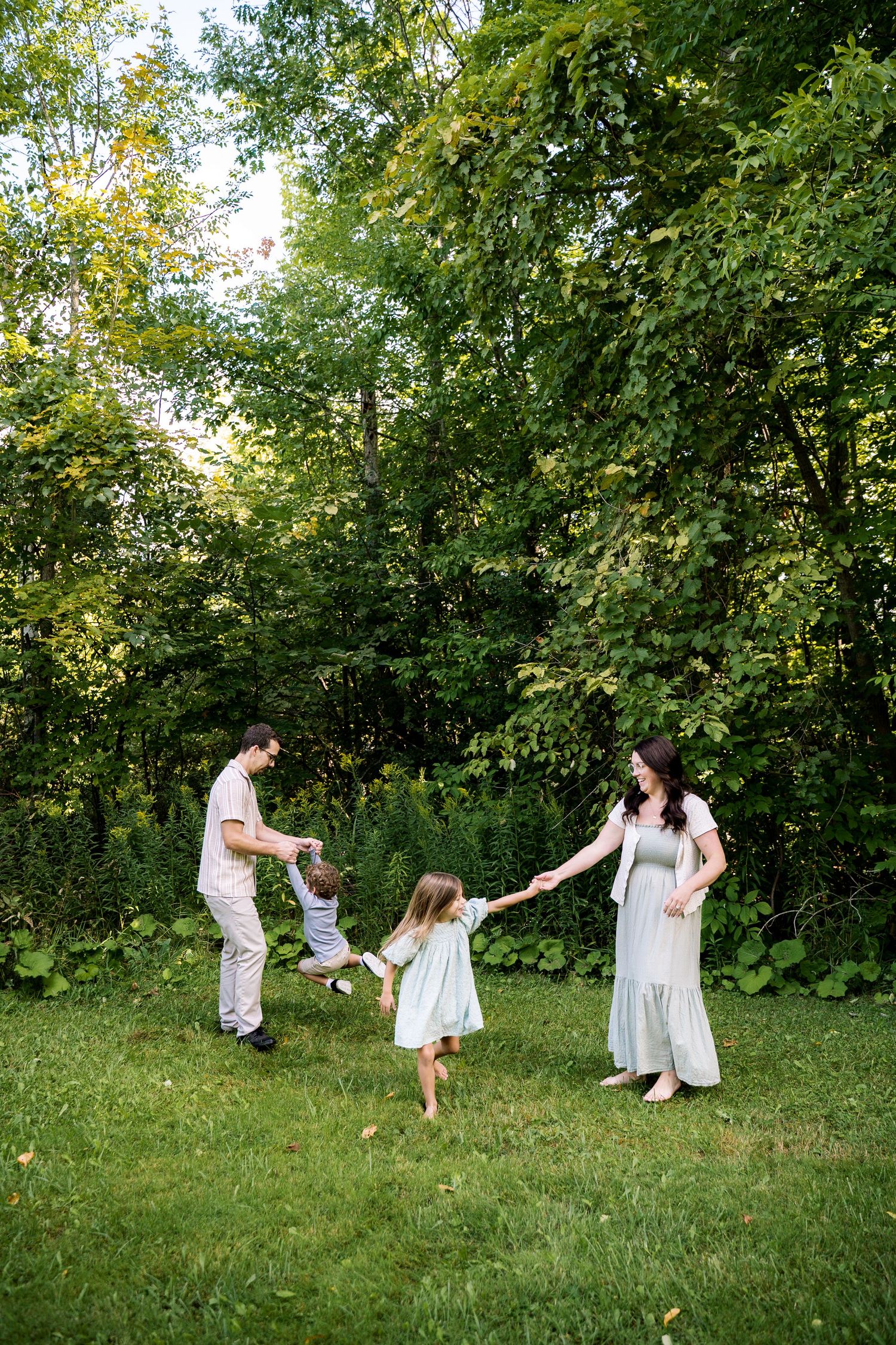 A family in white dresses dances and spins together on a grassy hill surrounded by lush green trees on a sunny day.