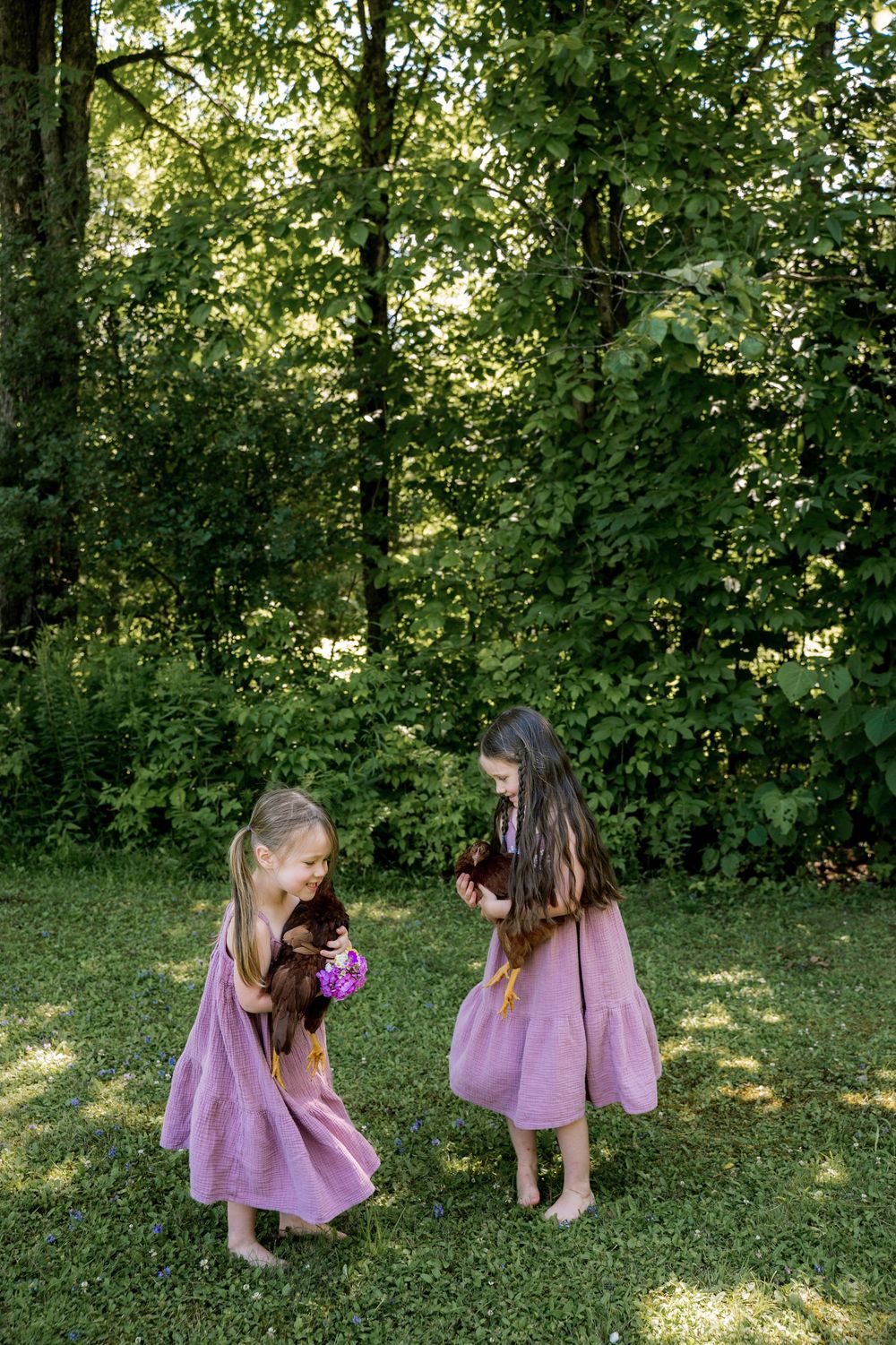 Two young children in purple dresses play together on a grassy lawn surrounded by lush greenery.