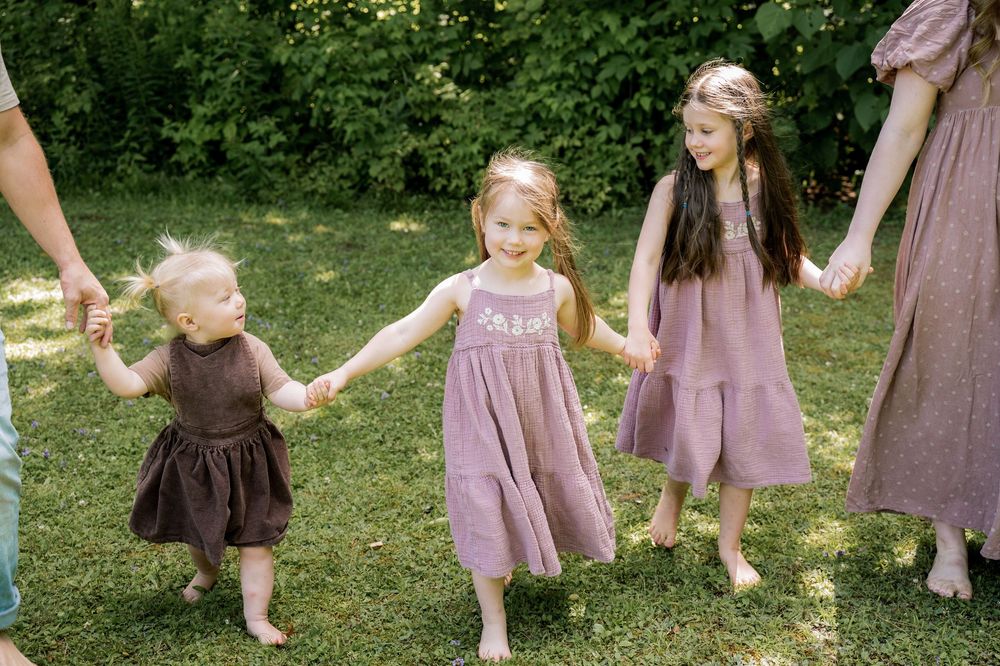 Children in purple and brown dresses playing and dancing together on grass in a sunlit outdoor setting.
