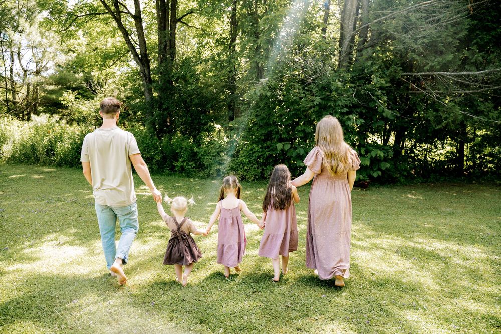 Family walking together through a sunlit garden path wearing coordinated purple and neutral outfits.
