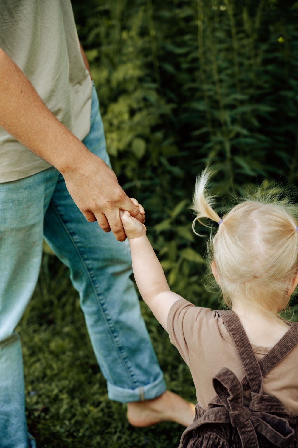 A sequence of four images showing a person helping a toddler learn to walk on grass in a garden setting.