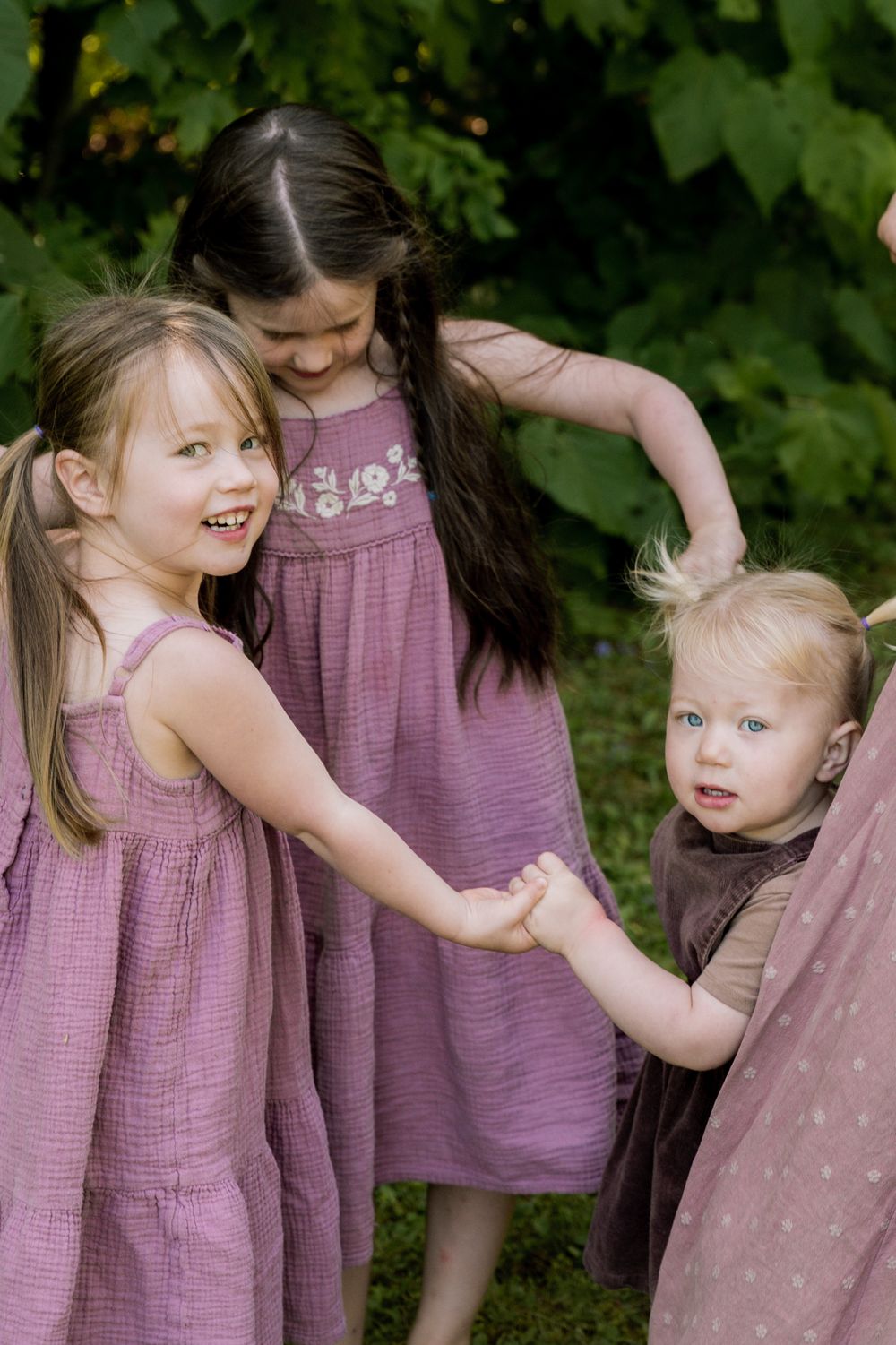 Children in matching purple dresses play and laugh together against a backdrop of green foliage.