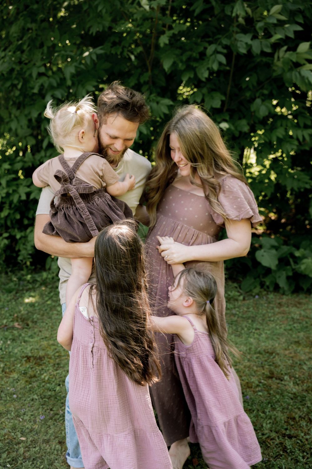 A family in coordinating mauve dresses and casual wear share an intimate moment together in a garden setting.