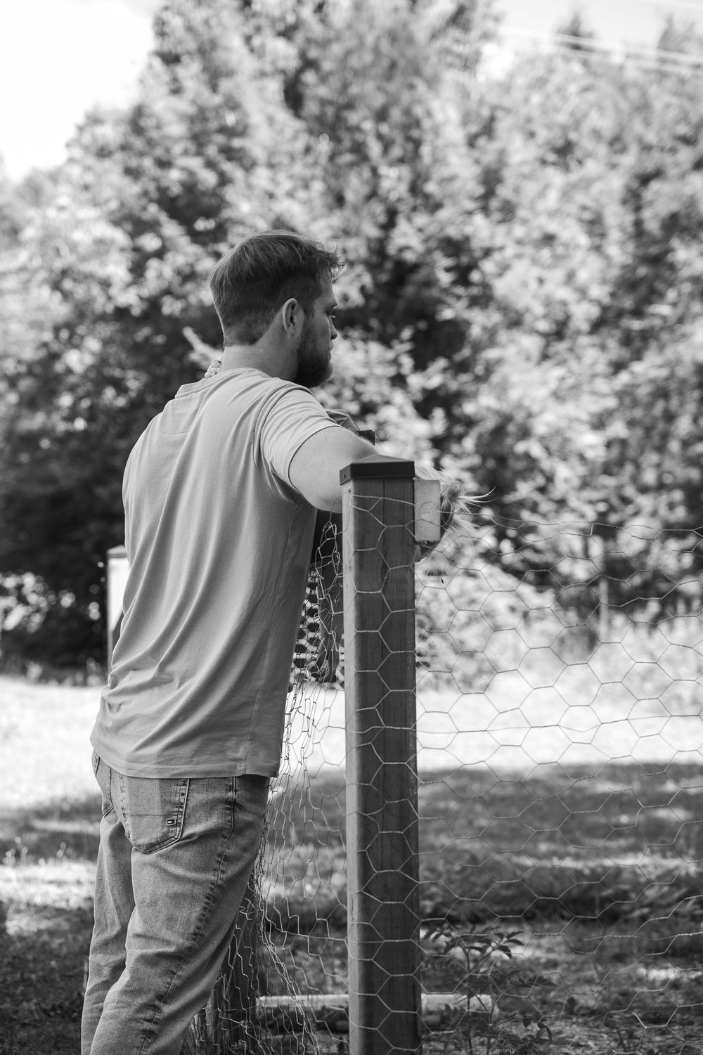 Black and white image of someone leaning against a wooden post in a tranquil outdoor setting.