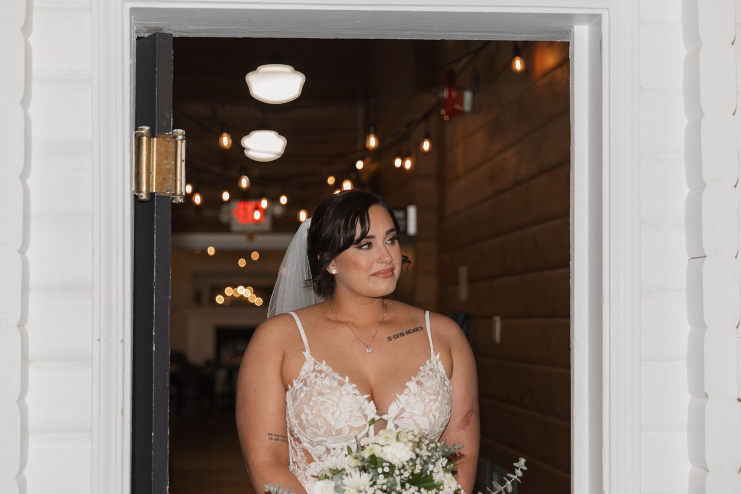 Beautiful bride in lace wedding gown holding white bouquet standing in rustic venue doorway.