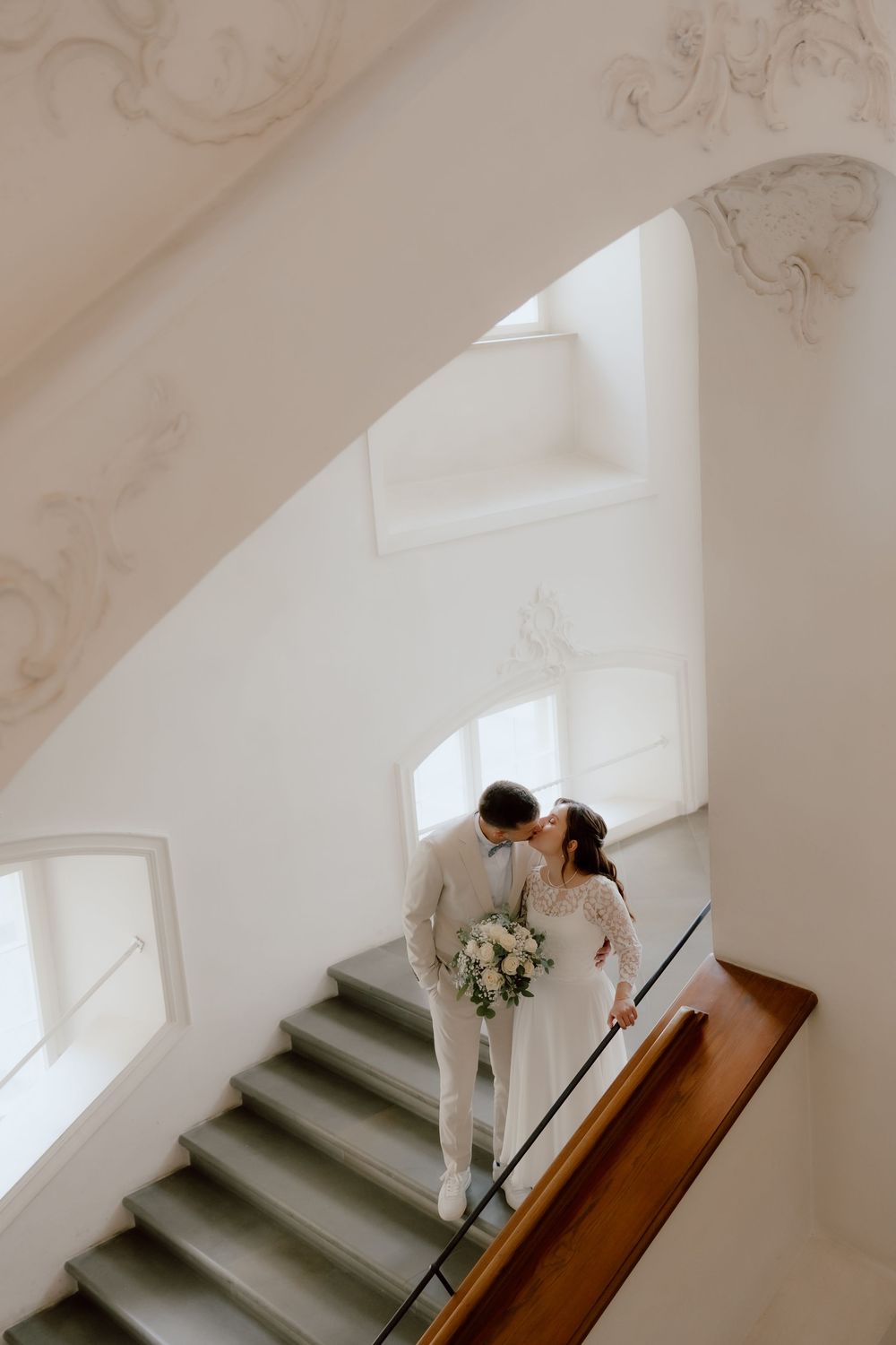 Elegant wedding couple descending white stairs in a historic building with ornate details.