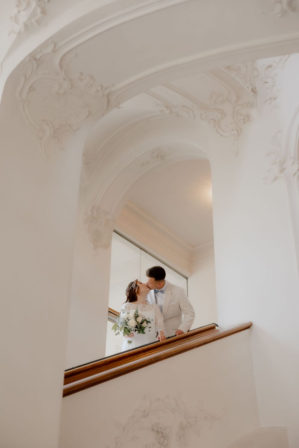 A romantic wedding couple shares an intimate moment in an elegant white archway overlooking a bright interior space.