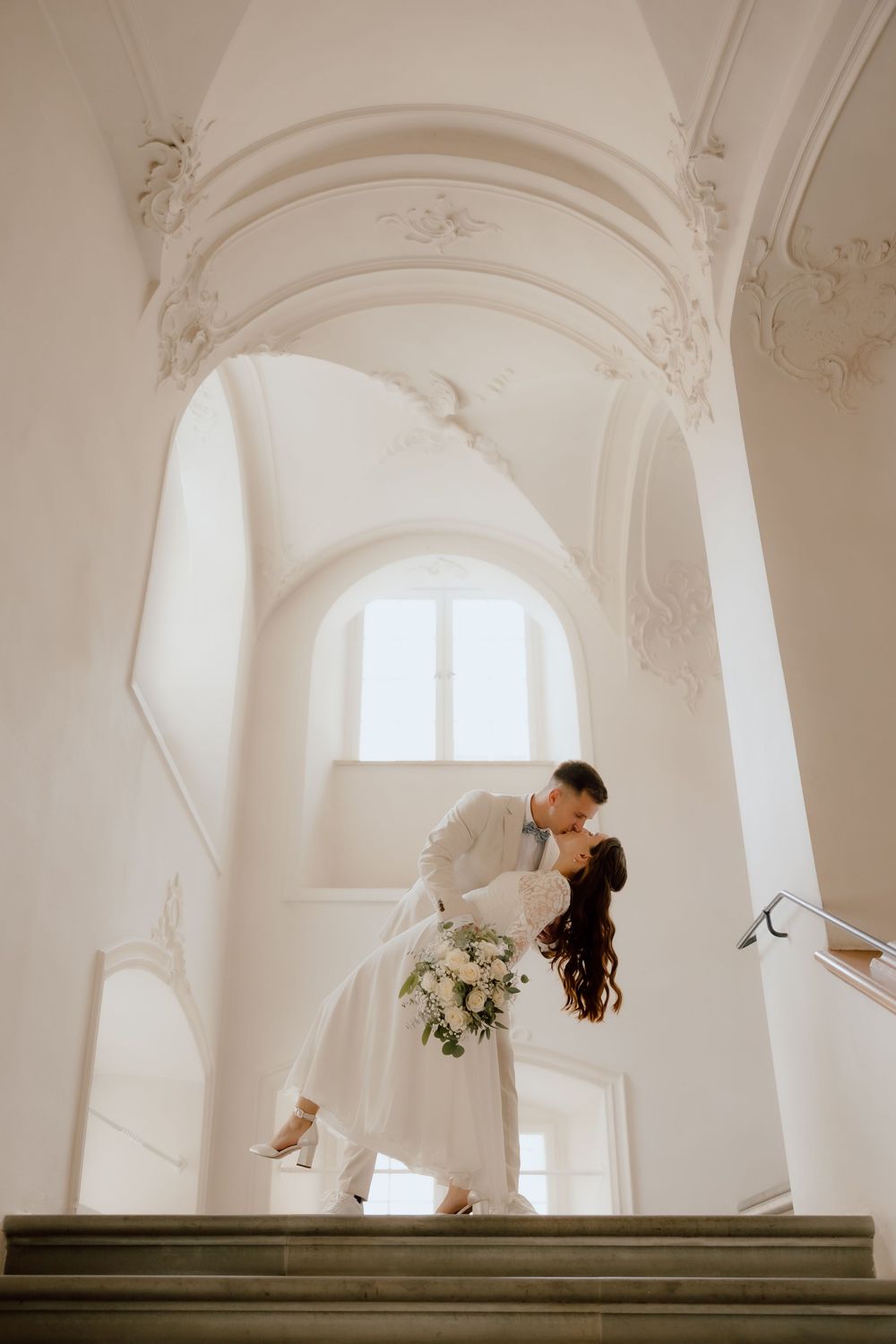 Romantic wedding couple dances and dips on elegant white stone staircase in historic building.