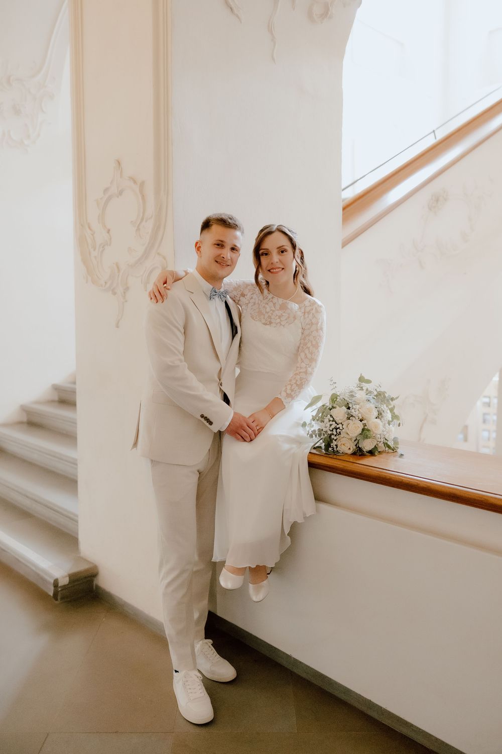 A couple in white attire poses together against a light-colored wall with a staircase in an elegant interior setting.