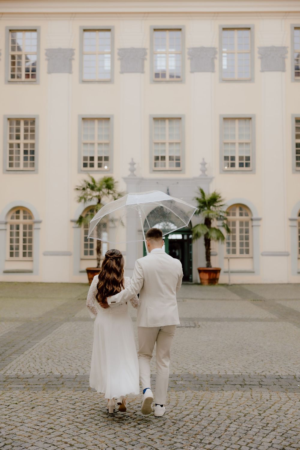 A couple in wedding attire walks along a cobblestone courtyard outside an elegant cream-colored building.