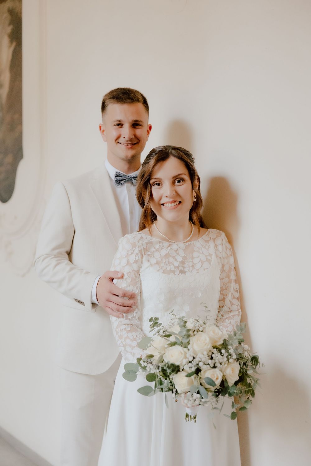 A bride and groom pose against a white wall, with the bride holding a delicate bouquet of white flowers and greenery.