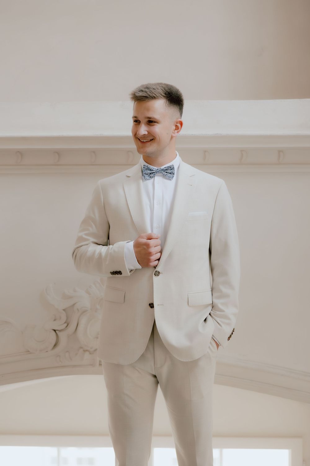 Groom in light colored suit poses in front of white wall at wedding venue.