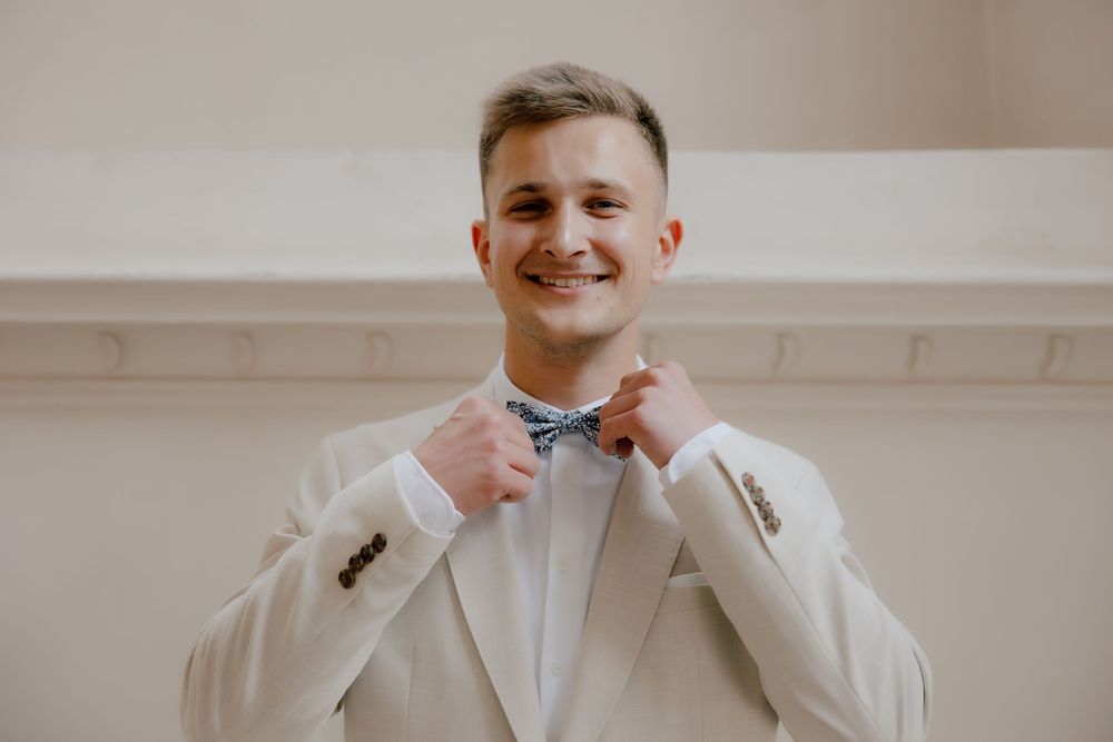 A person in a white suit and tie smiles while standing in front of a light colored wall.