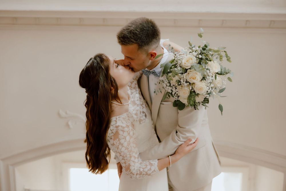 A romantic wedding moment captured in a bright indoor setting with elegant floral bouquet and lace dress details.
