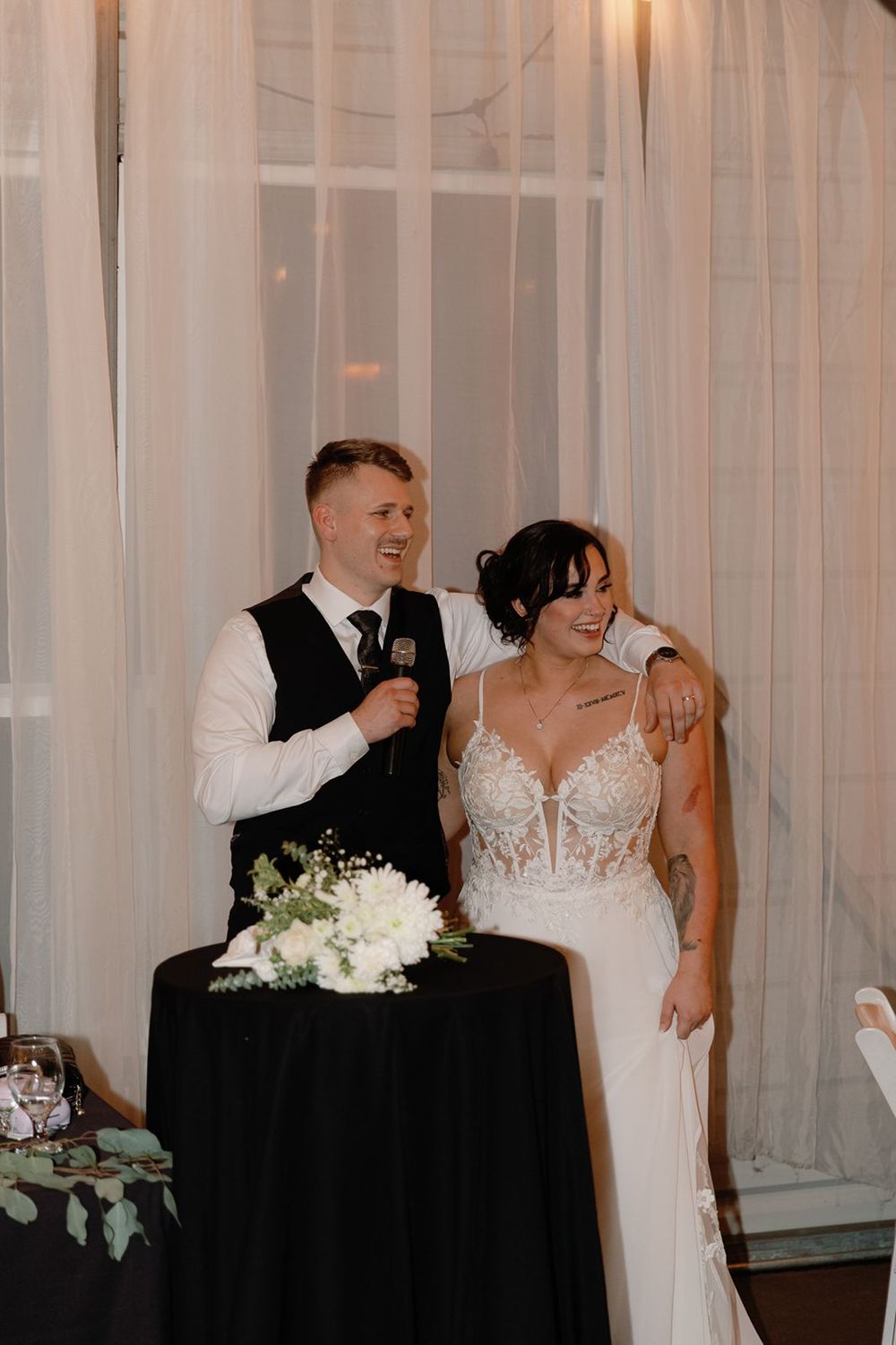 Bride and groom smiling together at sweetheart table with microphone during wedding reception speeches.