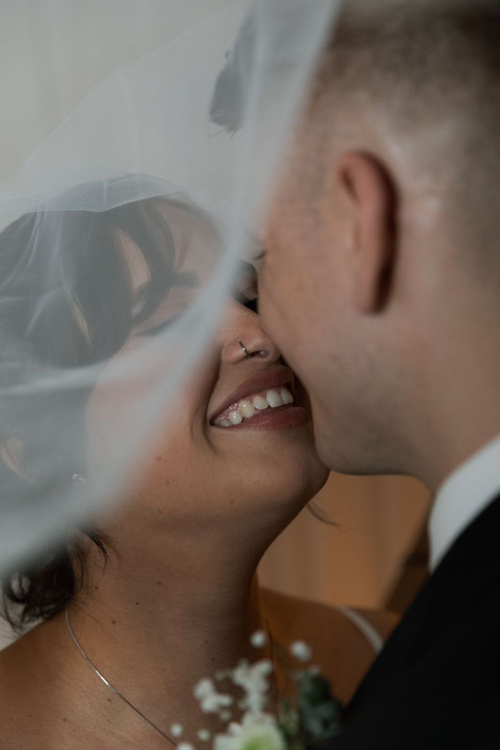 Bride smiling joyfully as groom kisses her cheek beneath flowing white veil.
