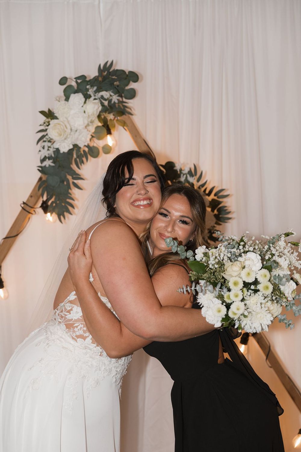 Bride in lace gown hugging bridesmaid in black dress before floral arch backdrop.