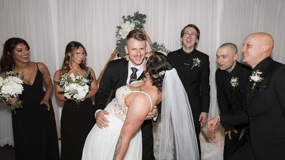 Bride and groom sharing first kiss surrounded by wedding party in black attire.
