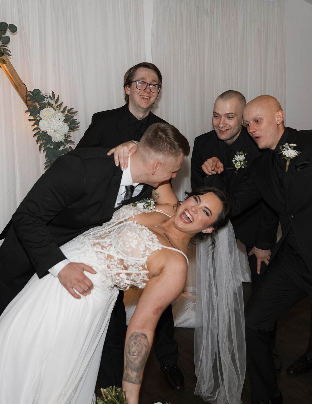 Groom dipping bride while groomsmen in black suits cheer and celebrate behind them.