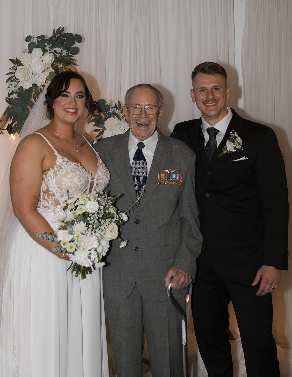 Bride and groom posing with elderly veteran wearing military medals at wedding reception.