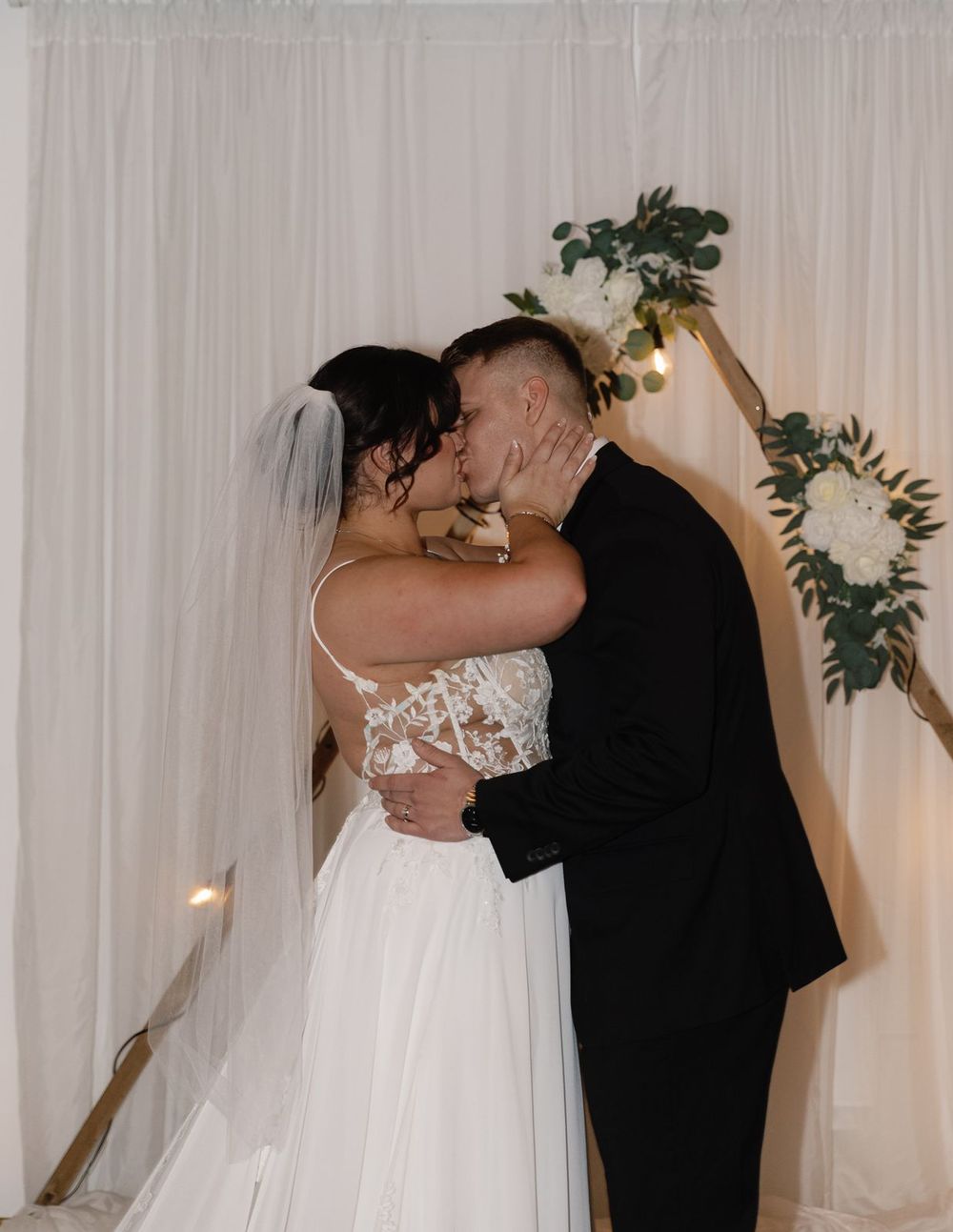 Bride and groom sharing first kiss at altar decorated with white flowers and greenery.