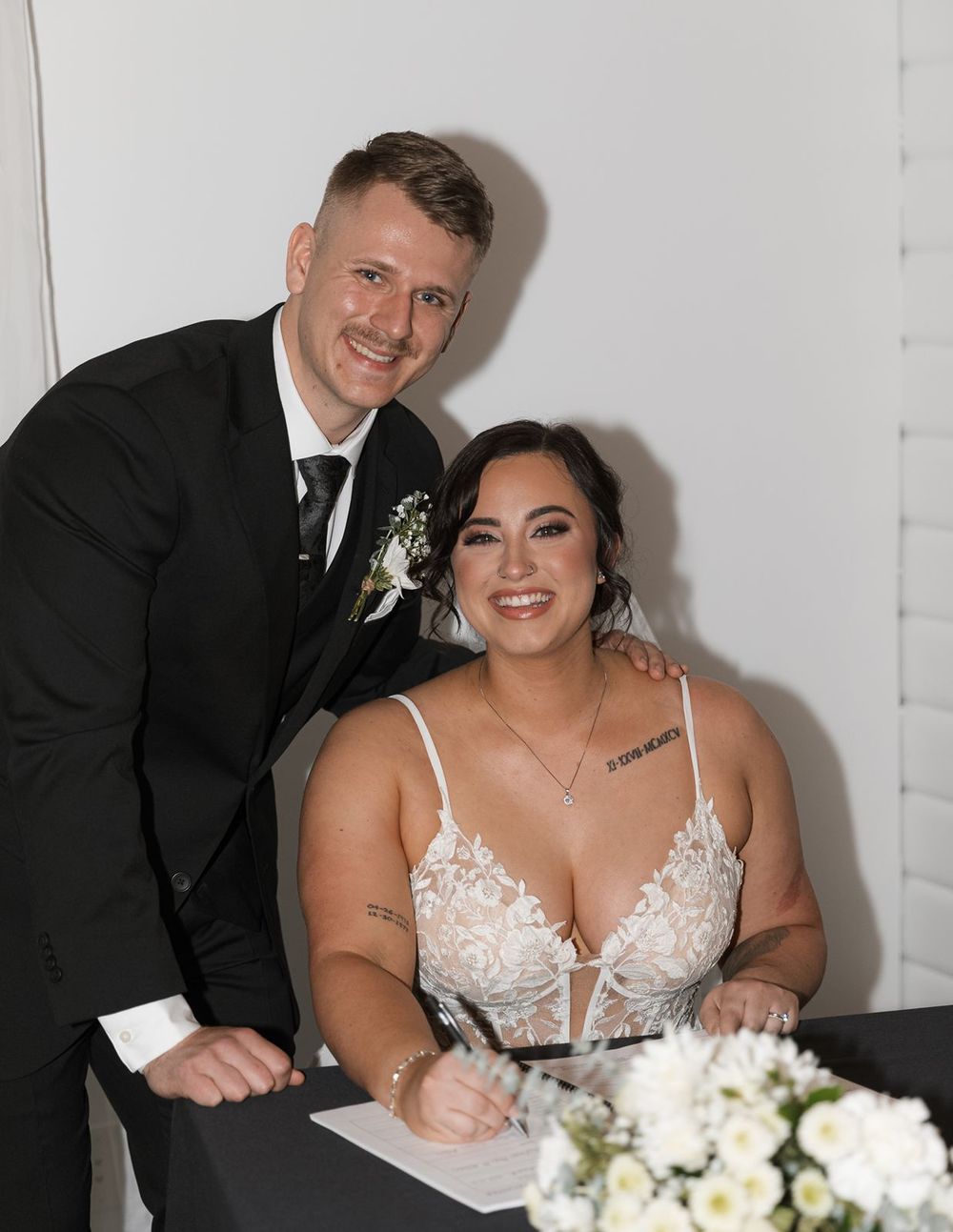 Bride signing marriage certificate while groom stands proudly beside her smiling.