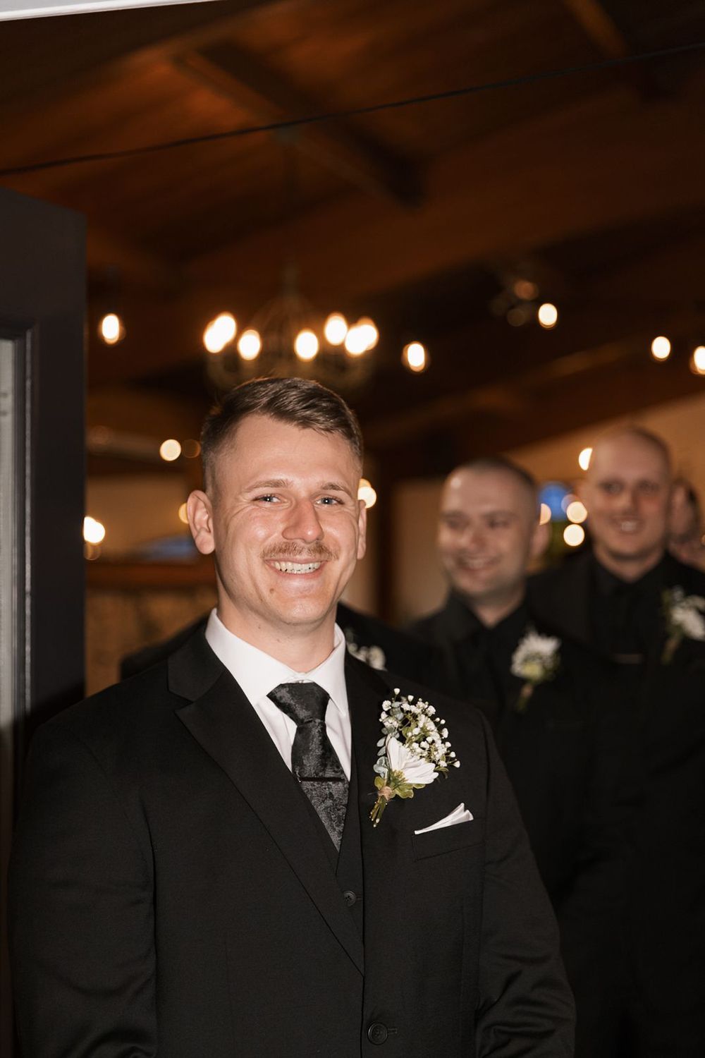 Smiling groom in black suit with boutonniere stands before groomsmen at elegant wedding venue with warm string lights.
